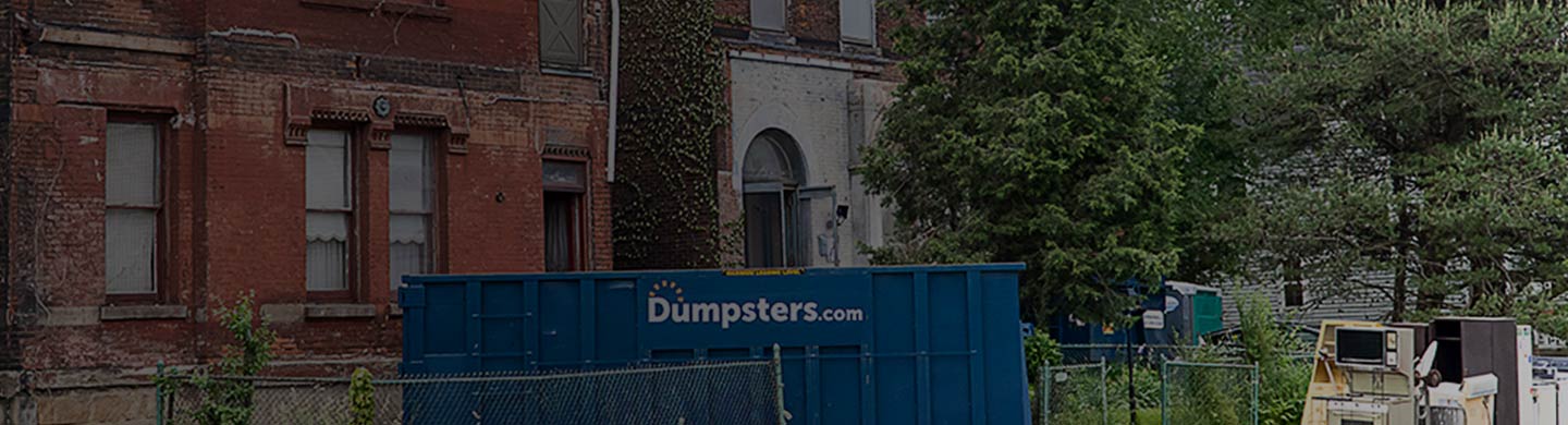 A Dumpsters.com Roll Off Dumpster Outside of a Brick Building.