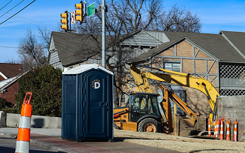 Dumpsters.com portable toilet on a construction site sitting next to a backhoe with an orange traffic cone in the foreground.