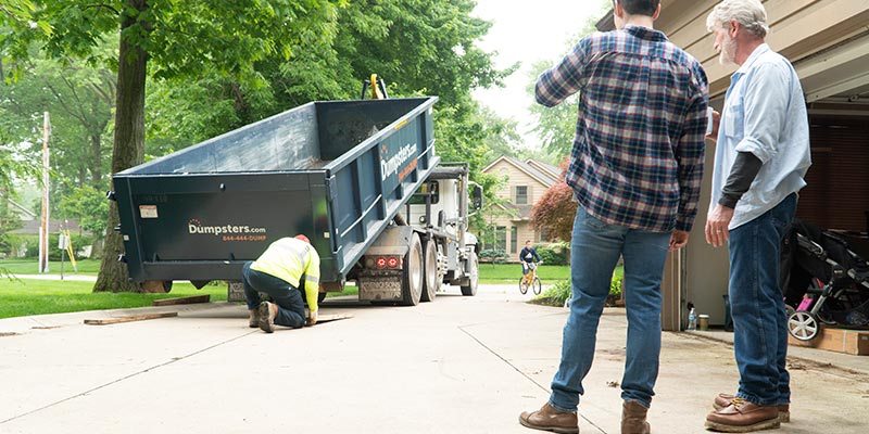 A Dumpsters.com Dumpster Truck Driver Puts Boards on a Driveway as Two Customers Watch.
