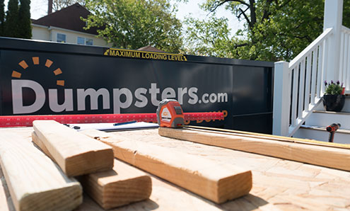 Blue Dumpster With Large Dumpsters.com Logo in the Background and a Pile of Wood Planks in the Foreground