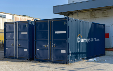 Two blue Dumpsters.com storage containers sitting side-by-side behind a building.